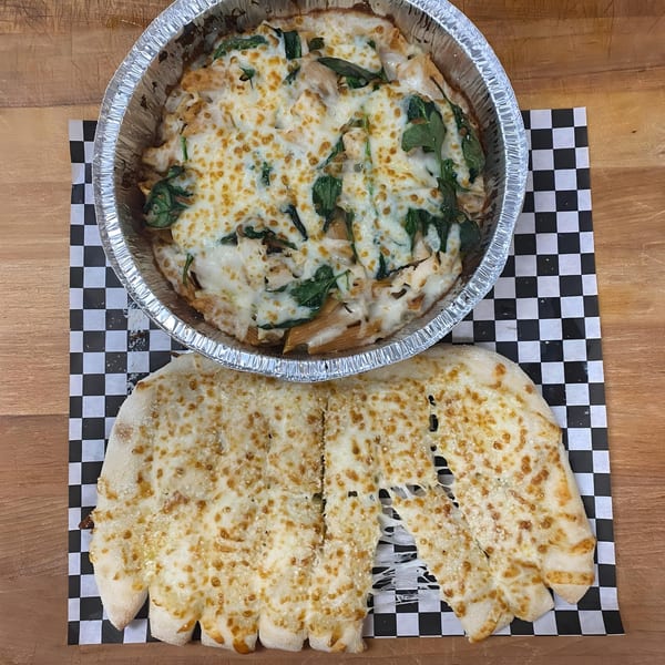 Baked pasta with melted cheese and spinach in a foil pan, with cheesy breadsticks on checkered paper.