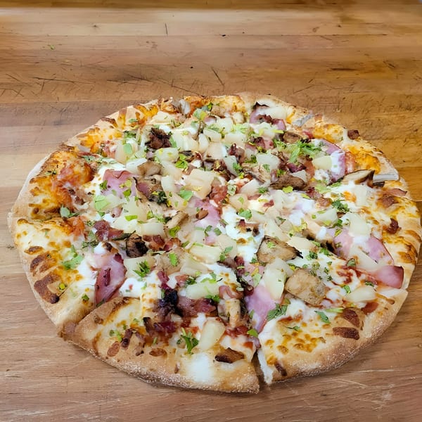 A whole No Name Pizza with pineapple, ham, bacon, chicken, and a white sauce on a wooden surface.